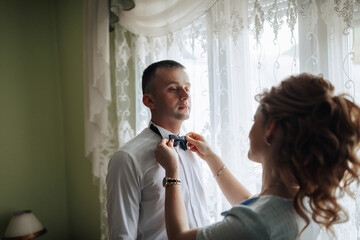 A bride and groom are standing in a living room, with the bride wearing a veil and the groom holding a bouquet