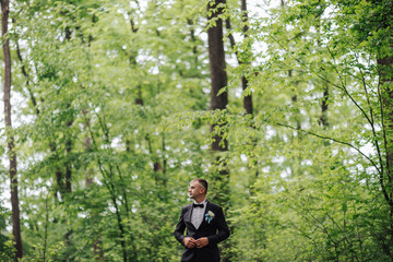 A man in a suit stands in a forest, looking at the camera. Scene is serene and peaceful, as the man is surrounded by nature