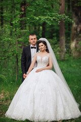 A bride and groom are standing in a forest, with the bride wearing a white dress and the groom wearing a black suit