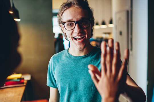 Overjoyed teen male hipster in spectacles ging high five to colleague celebrating success of completed project,cheerful young caucasian man waving to friend happy to see and greeting indoors