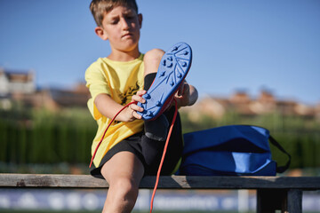 Young boy lacing up blue soccer cleats on a bench at the sports field