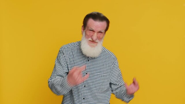 Overjoyed happy senior man showing rock n roll gesture by hands cool sign shouting yeah with crazy expression dancing rejoicing victory win success. Elderly bearded grandfather on yellow background