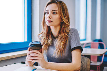 Thoughtful female student pondering on essay for university during coffee break indoors, young pensive woman holding takeaway cup sitting at desktop with notebook and thinking about education