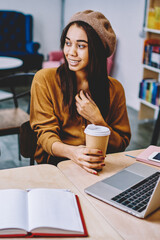 Positive successful hipster girls talking during coffee break after searching information for school project via laptop device, happy women enjoying caffeine beverage and friendly meeting indoors