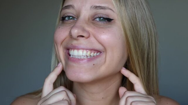 Young caucasian smiling blonde woman showing dimples on her cheeks on a dark grey background. Close-up of a blonde girl with even white teeth and beautiful smile