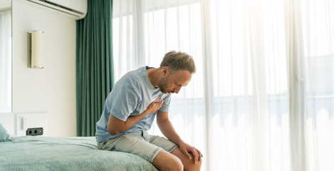 Middle-aged man sitting on bed, clutching chest in pain, heart attack or respiratory issue.