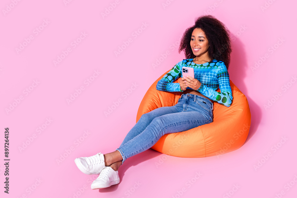 Poster Full size photo of lovely young lady sit orange beanbag hold device dressed stylish blue garment isolated on pink color background