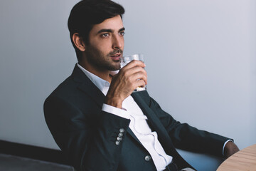 Pondering businessman drinking water in break of communication with colleagues via laptop application, indian man wear in formal suit thoughtful looking away holding glass sitting at desktop