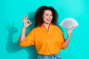 Photo of successful woman with wavy hairstyle dressed orange shirt hold money show okey wink eye isolated on teal color background