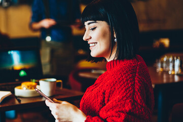 Cheerful brunette woman reading text message from friend using application for chatting, positive smiling hipster girl spending time for lunch at cafeteria and communicating with internet followers