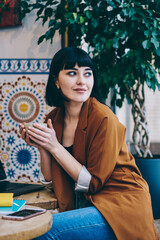 Smiling caucasian hipster girl holding cup with tea and looking at cafeteria window in break of working on laptop computer, modern netbook with mockup screen and textbook staying on wooden table