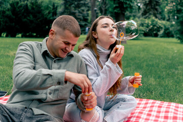 Obraz premium in a park in nature young couple sitting on a red checkered blanket blows soap bubbles back to childhood