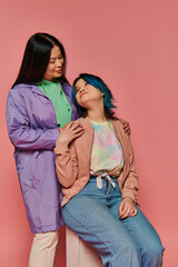 Two women, an Asian mother and her teenage daughter, stand together in casual wear against a vibrant pink background.