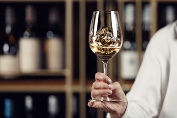 Glass of wine filled with white wine in female hand on cellar and bottles shelf background, close up with a free copy space for your text. 