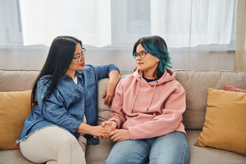 An Asian mother and her teenage daughter in casual wear engage in a deep conversation while sitting on a cozy couch.