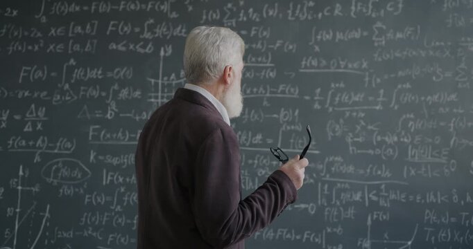 Pensive senior man professor looking at chalkboard with formula holding glasses thinking about science in university. Education and knowledge concept.