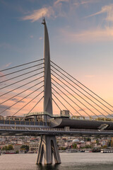 The Golden Horn Metro Bridge in Istanbul, Turkey, features a unique design with a tall, slender tower and a pedestrian walkway. The sunset casts a warm glow over the view