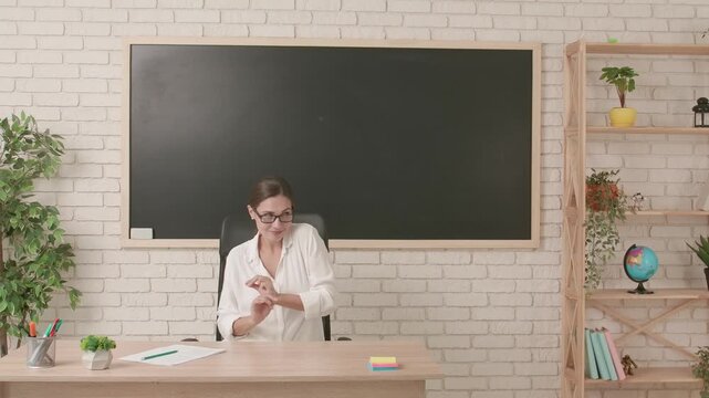 Woman college teacher sitting at desk in classroom in front of chalkboard and doing little funny dance smiling at camera. Education concept.