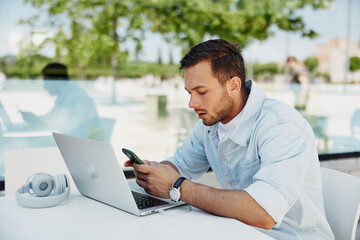 Young man focused on smartphone while working on a laptop in a modern outdoor café