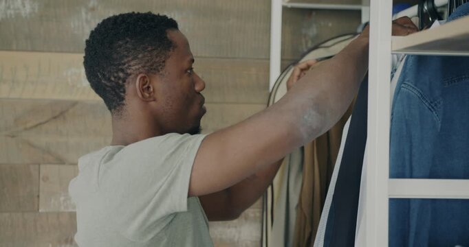 Attractive African American man choosing stylish clothes on hanger and shelf in wardrobe looking for fashionable outfit at home. People and style concept.