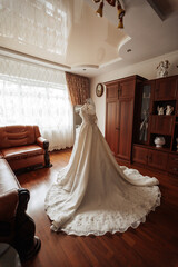 A bride's wedding dress is displayed in a room with a wooden floor and a brown couch. The dress is white and has a long train