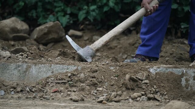 Worker using chisel and hammer to dig in construction site. Construction worker in action, using a chisel and hammer for digging and breaking the ground at a construction site surrounded by greenery.