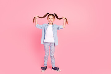 Full size photo of good mood adorable small girl holding long ponytails dressed denim shirt having fun isolated on pink color background