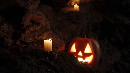 Jack-O'-Lantern Pumpkin in a Dark Cave, Wind Blowing Out Candles on the Rocks. Creepy Atmosphere of an Approaching Spirit.