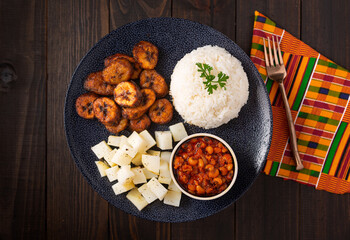 West African dish of fried plantains, cubed potatoes, brown peas and white rice on a wooden surface with a kente cloth napkin; copy space; directly above