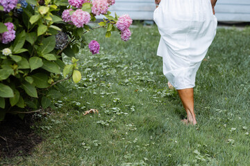 torso of woman in white flowing summer dress walking through lush backyard next to a purple hydrangea bush