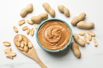 Tasty peanut butter in bowl and spoon with groundnuts on white table, flat lay