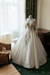 Fototapeta premium A white wedding dress is displayed on a mannequin in a room with a window. The dress is long and has a lace-like design