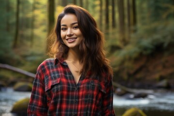 Fototapeta premium Portrait of a smiling asian woman in her 20s wearing a comfy flannel shirt in front of tranquil forest stream