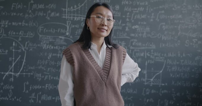 Zoom-in slow motion portrait of cheerful Asian lady school teacher standing in classroom with chalkboard smiling looking at camera. Education and professional occupation concept.