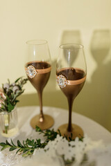 Two wine glasses with gold stems and a blue crystal bead on the stem. The glasses are on a table with a green leafy decoration