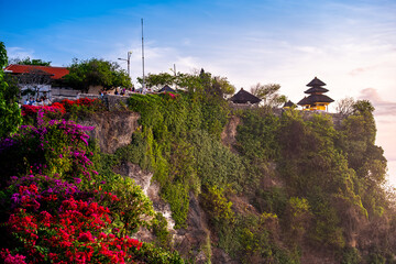 A temple on a cliffside, surrounded by lush greenery and vibrant red and purple flowers.