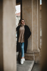 Obraz premium A woman dressed in a sleek black coat, white shoes, and blue jeans poses elegantly by an urban pillar. Perfect for showcasing contemporary fashion and urban lifestyle