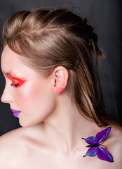 Naklejka premium Profile View of Woman with Butterfly on Shoulder. Young Caucasian woman with vibrant red and purple makeup, butterfly on shoulder, profile view against dark background. Vertical photo. Close-up.
