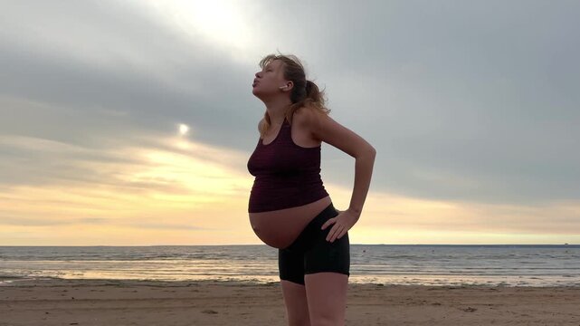 Young pregnant active woman doing exersice outdoors, stratching her body and meditate on the sea, beach at sunset in the evening