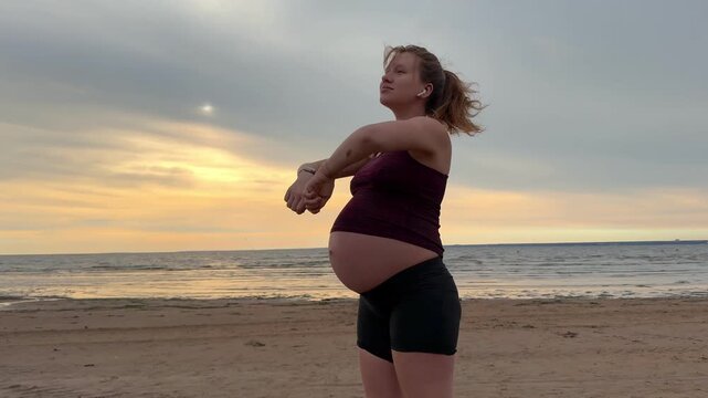 Young pregnant active woman doing exersice outdoors, stratching her body and meditate on the sea, beach at sunset in the evening