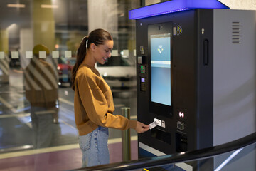 Young woman driver paying for ticket in parking meter near terminal in the underground parking area