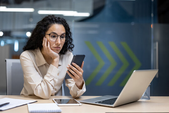 Bored office worker looking at phone feeling unproductive at desk with laptop, tablet, notebook in modern office environment. Represents lack of motivation and engagement.