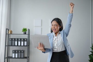 Businesswoman is holding a laptop and raising her arm in the air with joy, expressing her...
