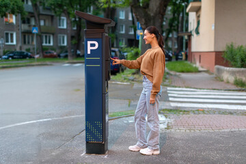 Young woman paying for parking in automated parking meter on the street, pressing buttons and...