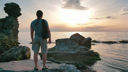 A lone individual stands by the water’s edge, gazing at a serene sunset, with rock formations...