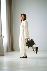 woman in a white lace dress and black handbag stands gracefully in a minimalist room. Her poised demeanor and elegant attire create a serene and sophisticated portrait.