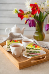Wooden tray with a cup of tea, a layered cake slice, and sandwiches; flowers in the background
