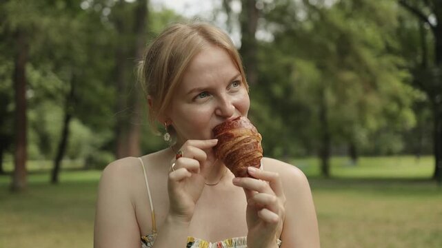 Young blonde Caucasian felame in trendy summer sundress eating croissants at a cafe at park. Girl bite piece of croissant look joyful. Carefree woman making picnic outside.. High quality FullHD