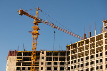 Towering yellow crane at a construction site against a clear blue sky, with a building under construction and visible scaffolding