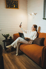 Obraz premium A woman in home clothes is sitting at home on the sofa with a laptop and talking on the phone. Work from home.